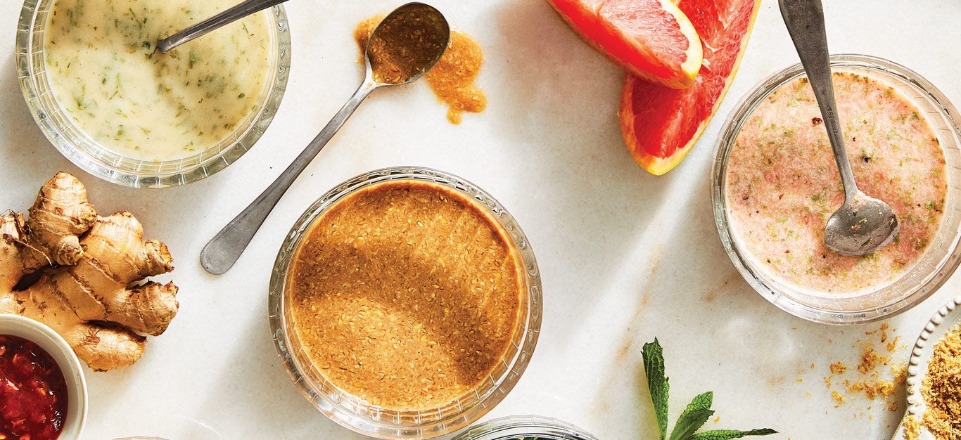 An array of homemade salad dressings made without oil, including vegan ranch, balsamic vinaigrette, and a grapefruit tarragon, in small glass bowls with spoons in them and some of the ingredients shown beside them
