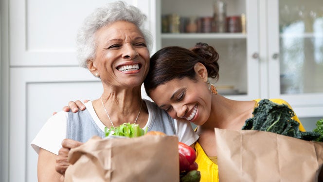 Mother and daughter lean on each other, smiling, while holding paper grocery bags full of veggies