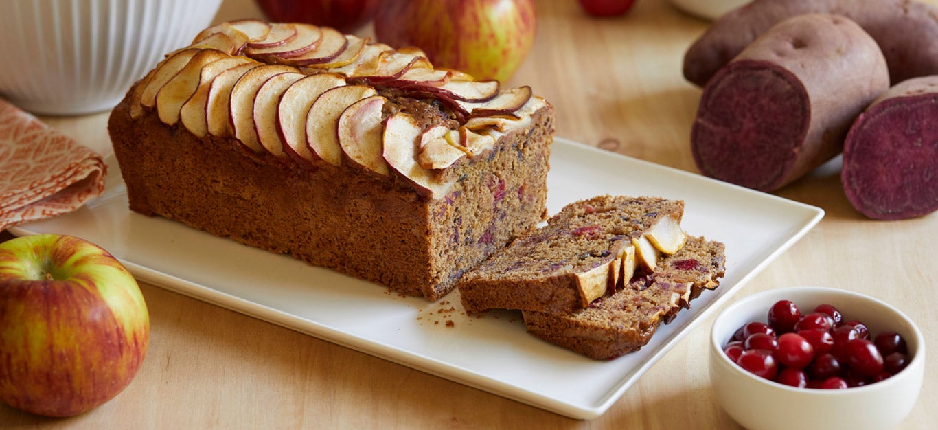 A sliced loaf cake with slices of baked apple arranged beautifully on top and a ramekin of cranberries to the side