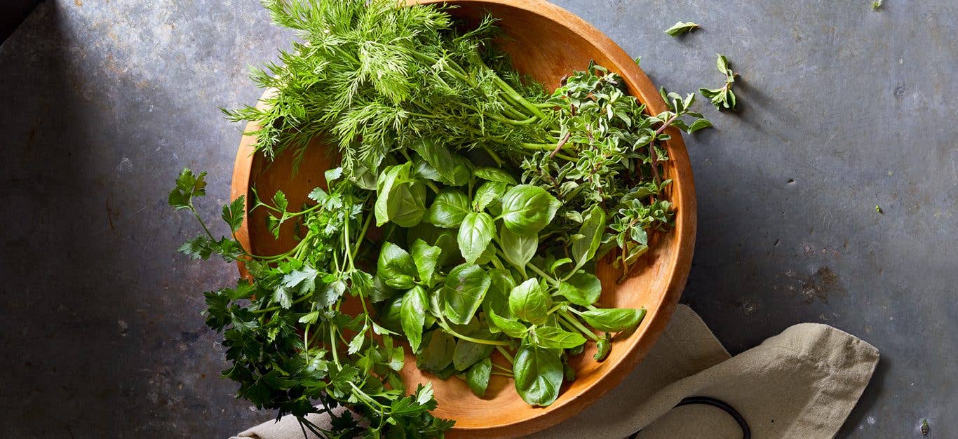 A bowl of snipped fresh green herbs, including mint, basil, oregano, and chives, sitting in a terra cotta bowl on a gray table