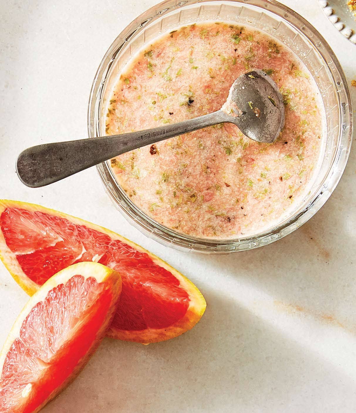 A light-pink homemade oil-free dressing with grapefruit and tarragon shown above in a clear glass cup or jar, with a silver spoon resting in it, with two wedges of grapefruit beside it
