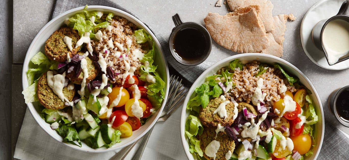 Two bowls loaded with a colorful mix of chopped lettuce, cucumbers, tomatoes over a pile of bulgur grains, topped with air-fryer falafel and drizzled with sauce