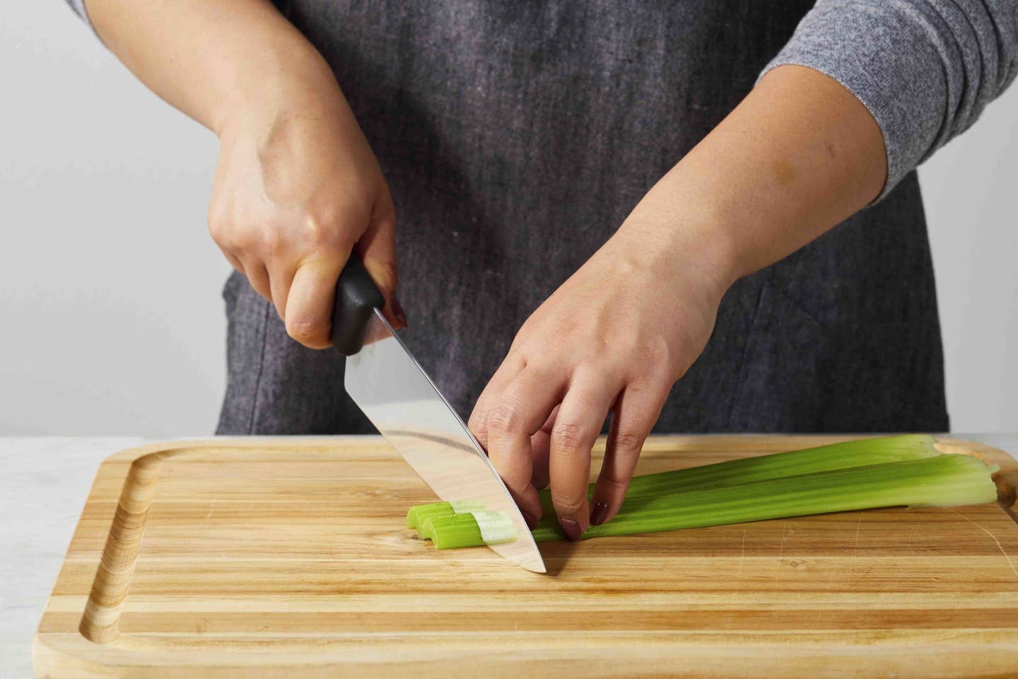 the midsection of a person standing behind a counter with celery on a cutting board, slicing the celery with a knife, demonstrating slicing techniques