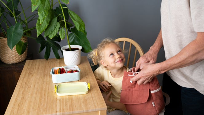 a little girl sits at a kitchen table as her did packs her lunchbox full of healthy foods, including strawberries and carrots