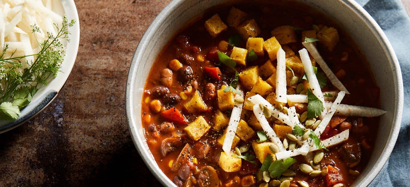 A pot of deep red mole-style vegan chili topped with corn and strips of jicama