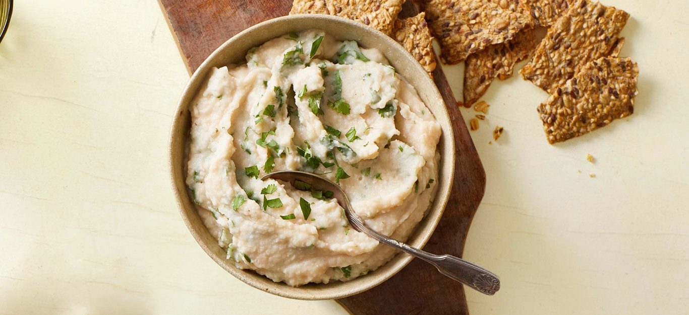 Navy Bean Homemade Hummus - white creamy hummus in a small bowl, garnished with cilantro, with whole grain crackers on the side