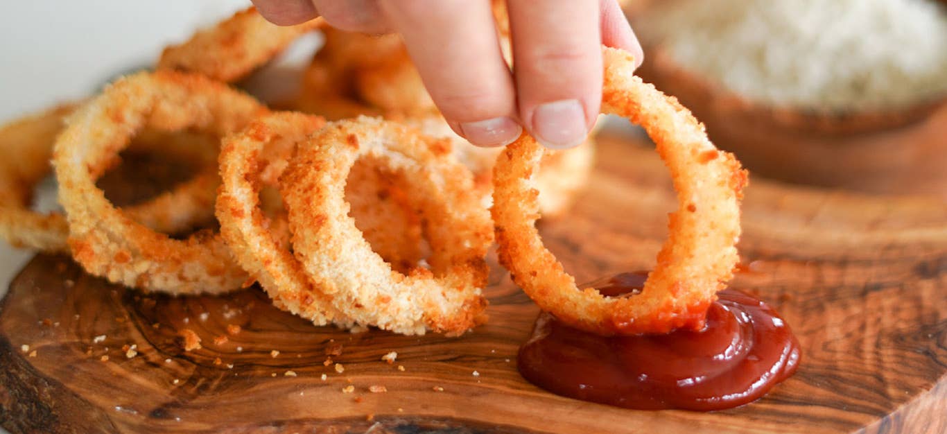 A photo of baked or air-fryer onion rings on a wooden tray, with a hand holding one ring and dipping it in ketchup