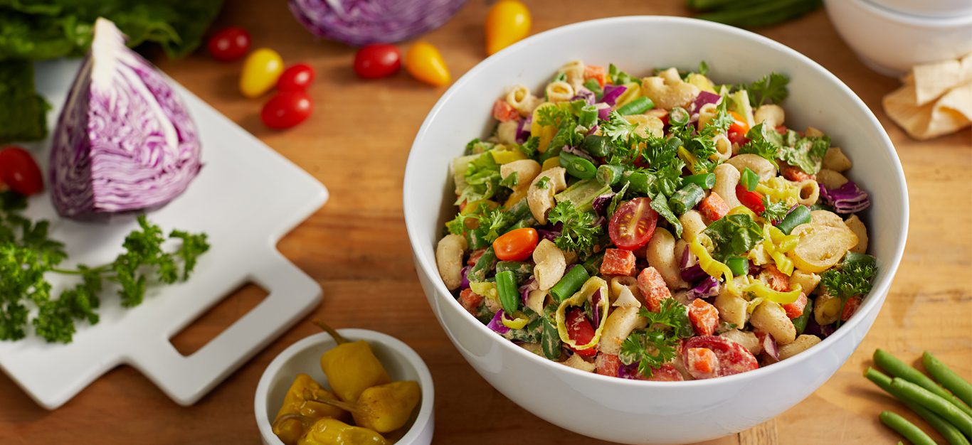 chopped veggie pasta salad in a white bowl next to a cutting board with chopped cabbage and herbs