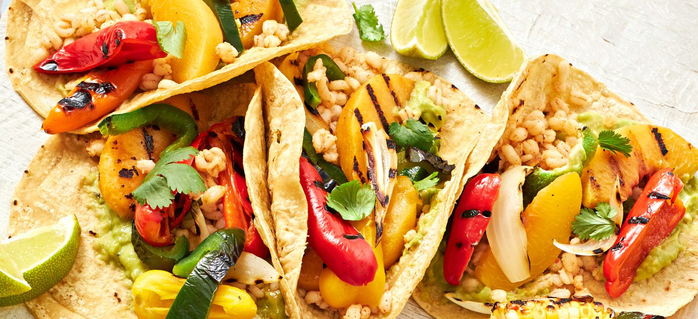 a close-up of four veggie tacos filled with peppers and grilled peaches, with lime slices on the side