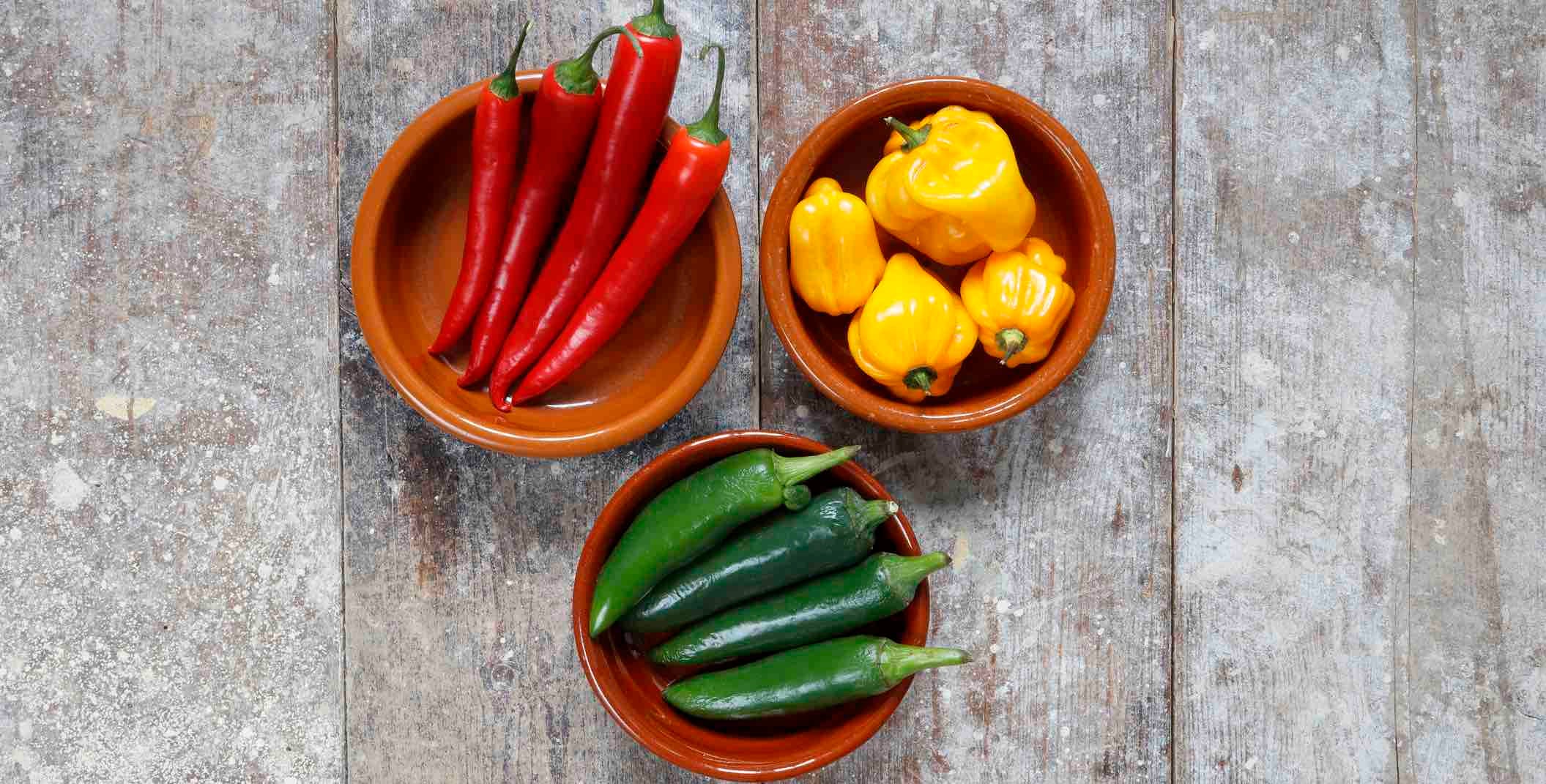 Three bowls, each containing a different type of fresh chile pepper, including thai bird, jalapeno, and scotch bonnet chiles, on a light gray wooden floor
