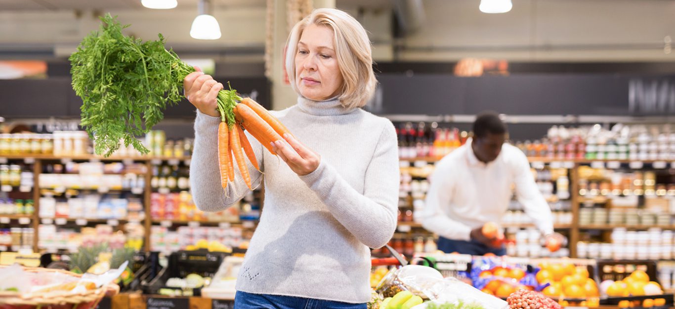 Middle-aged woman examines a big bunch of carrots in a grocery store produce section - Plant proteins have been shown to reduce risk of death in post-menopausal women