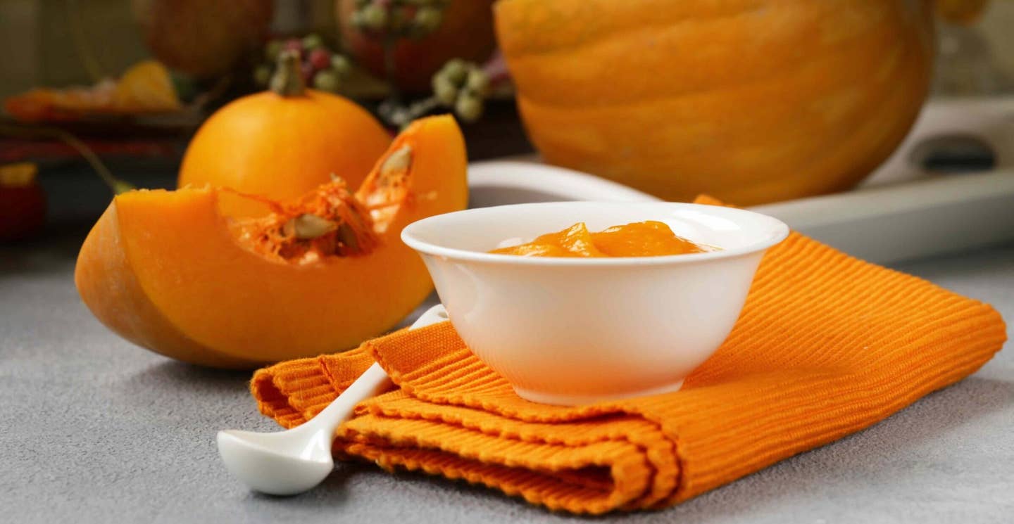 A bowl of pumpkin puree sits atop a folded orange kitchen towel, with cut pumpkin in the background