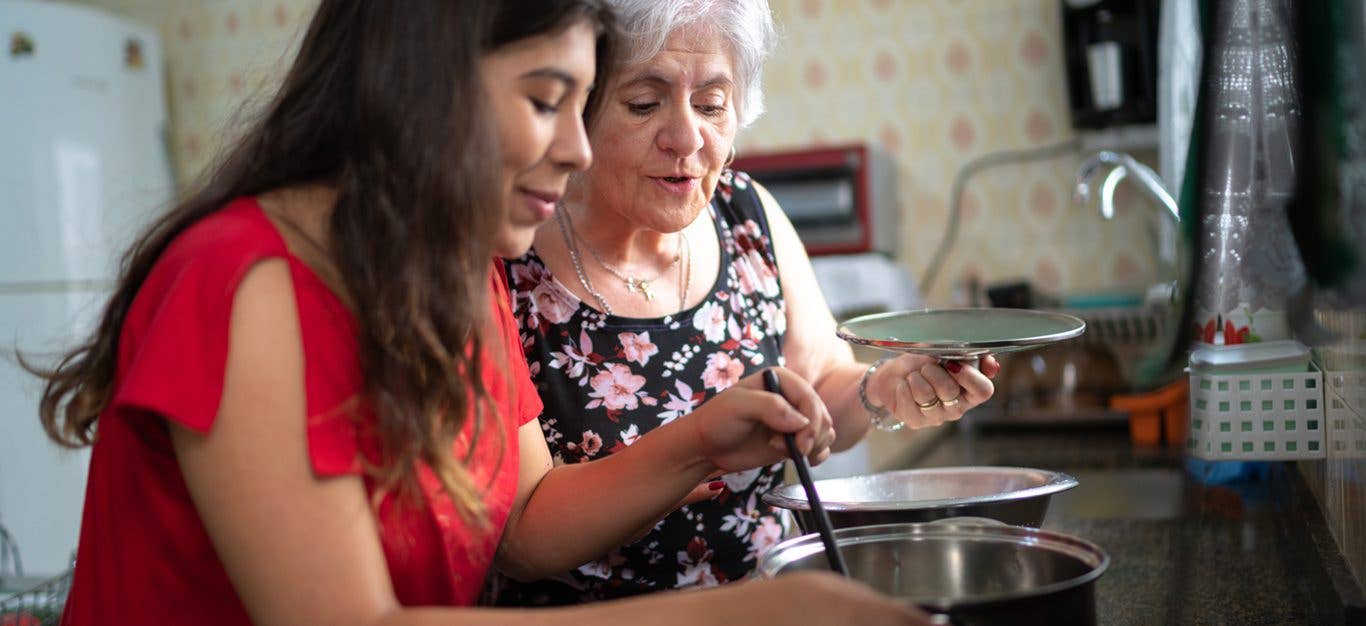 senior woman stands next to adult daughter or granddaughter with both women looking over a pot on a stove while smiling