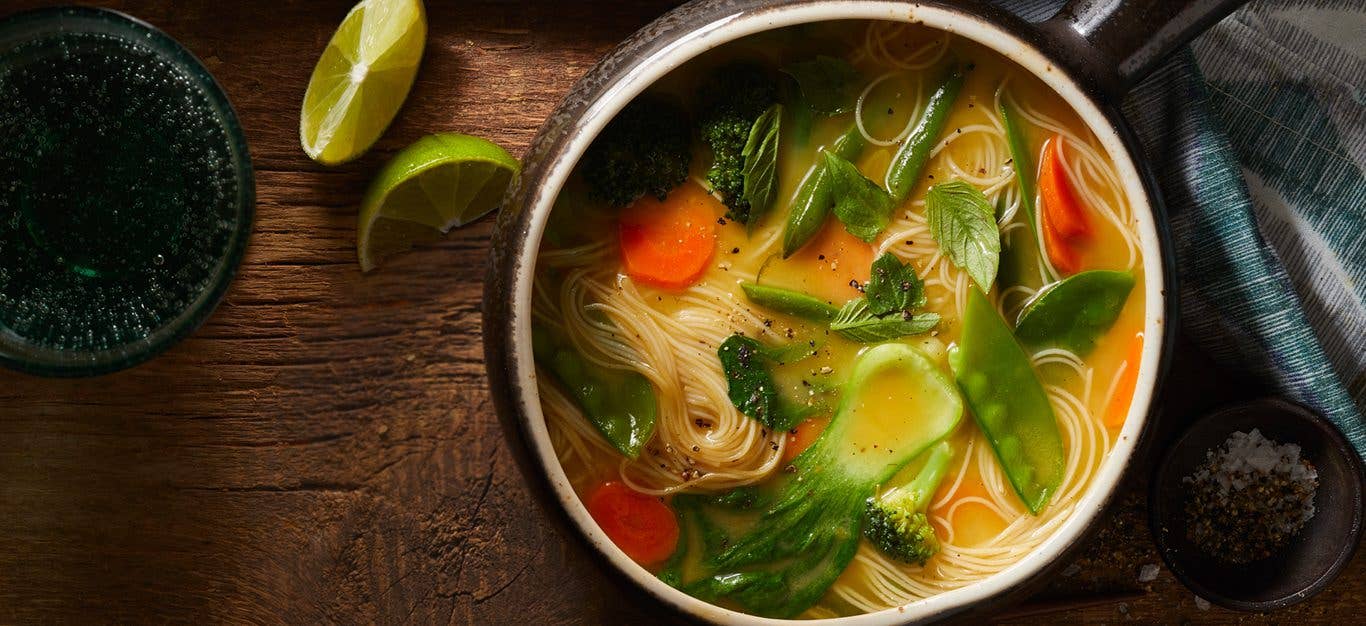 Vegan Thai noodle soup in a rustic pot, with noodles threading through slices of bok choy, carrots, broccoli, and snap peas, garnished with Thai basil, on a wooden table