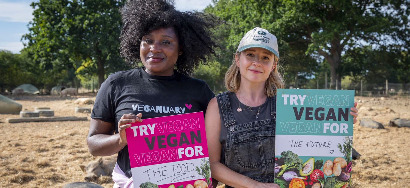 Two women standing in an animal sanctuary holding signs that say Try vegan for the food, and Try vegan for the future