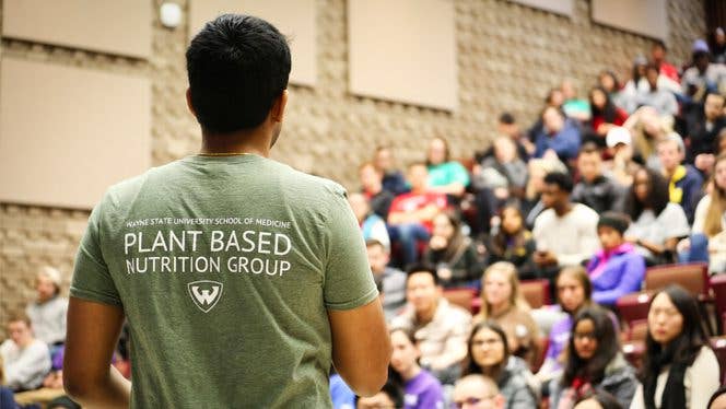 a man with a shirt that says "plant based nutrition group" speaks to a lecture hall of medical students