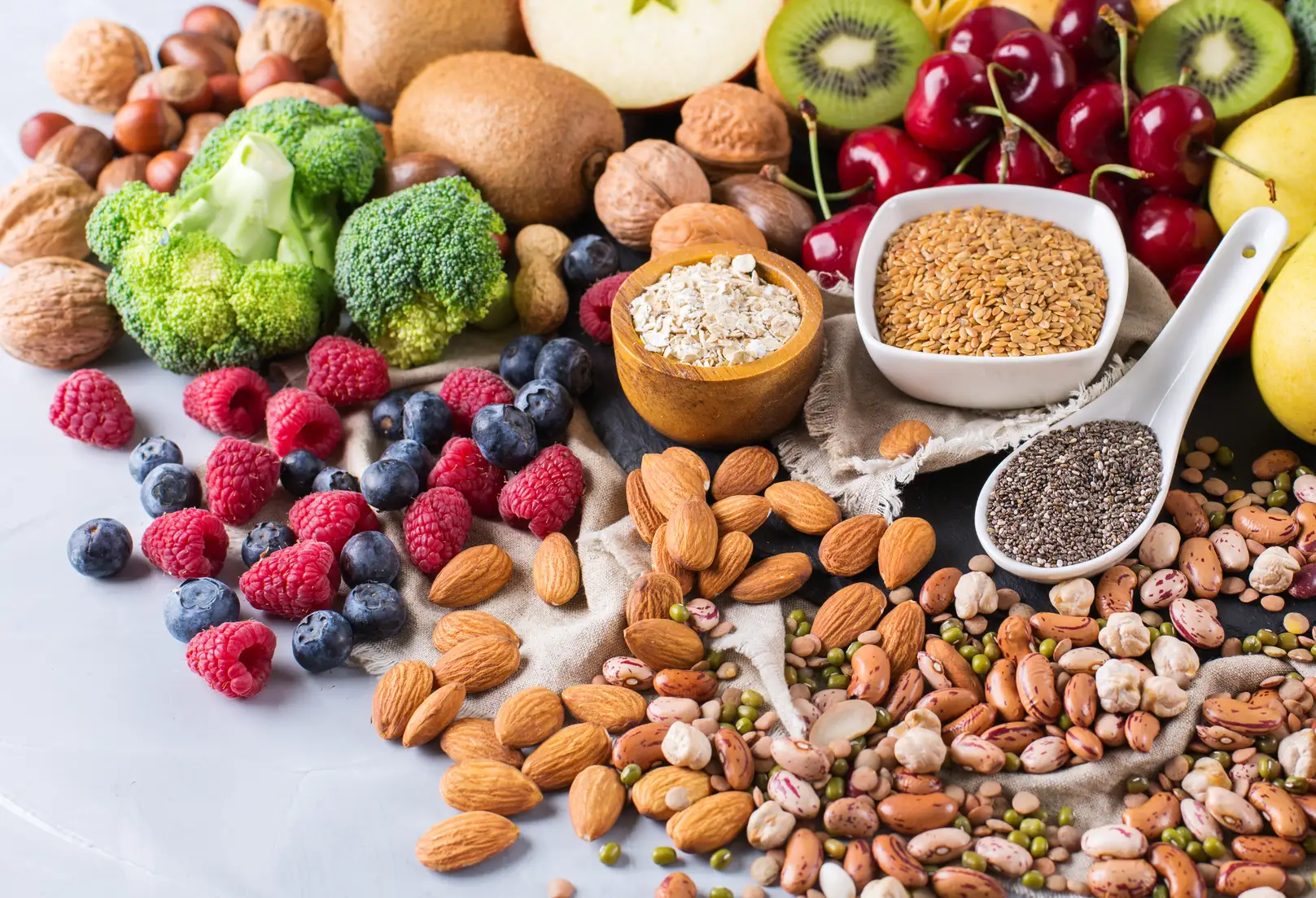 Assorted whole plant foods rich in antioxidants, including broccoli florets, berries (raspberries, blueberries, cherries), kiwi, apples, oats, flaxseeds, chia seeds, almonds, walnuts, and a variety of dried and cooked beans, arranged on a light surface.
