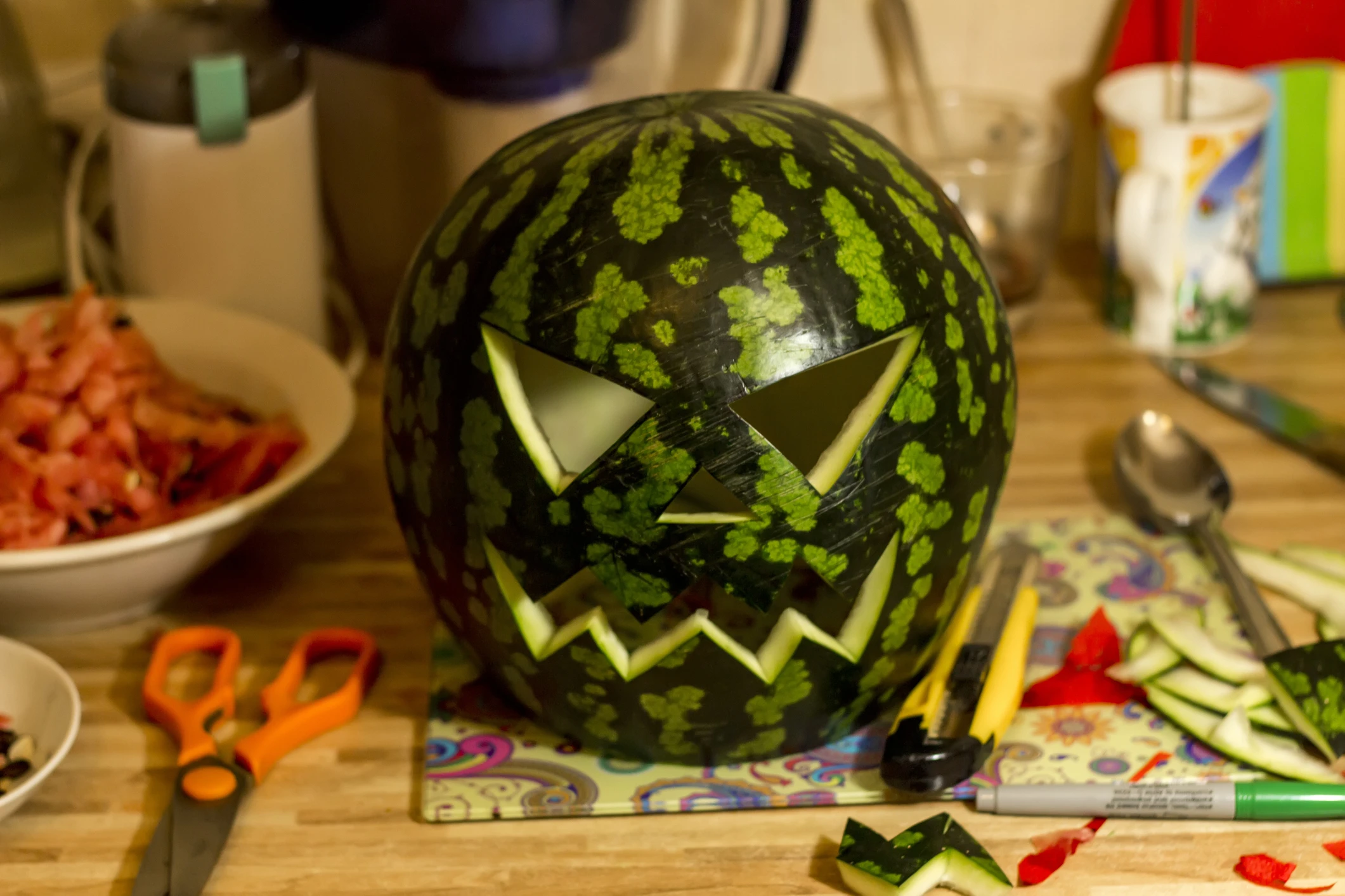 A watermelon with a face cut into it like a jack-o-lantern, surrounded by crafting supplies on a kitchen counter as a Halloween party food idea