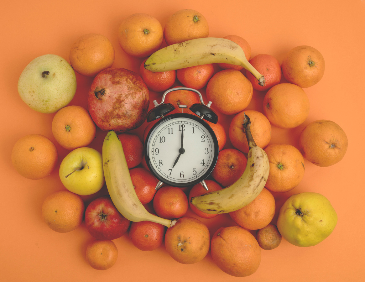 Alarm clock surrounded by orange and yellow fruits such as bananas and apples and oranges on an orange background