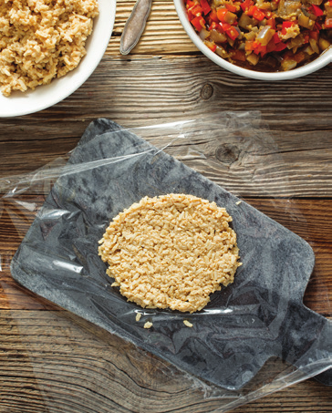 Photo illustrating the following step for making brown rice onigiri: Place an 8-inch square sheet of plastic wrap on the cutting board; place ¼ cup of rice in the center. Lift one end of the plastic to cover the rice and press down with your palm to flatten it, compacting the grains as tightly as possible.