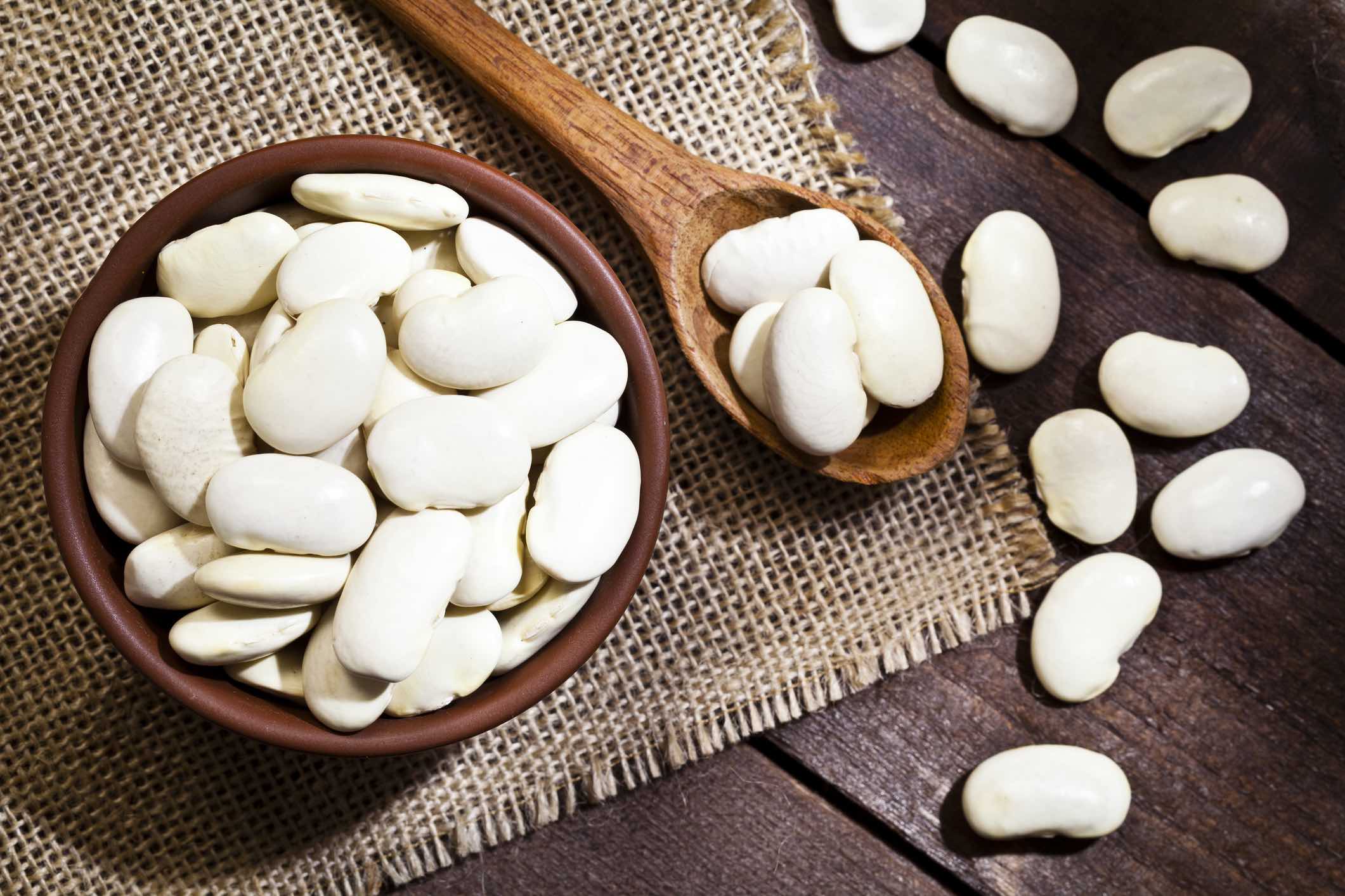 Top view of a brown bowl filled with lima beans shot on rustic wood table. A wooden spoon is beside the bowl with some beans on it.