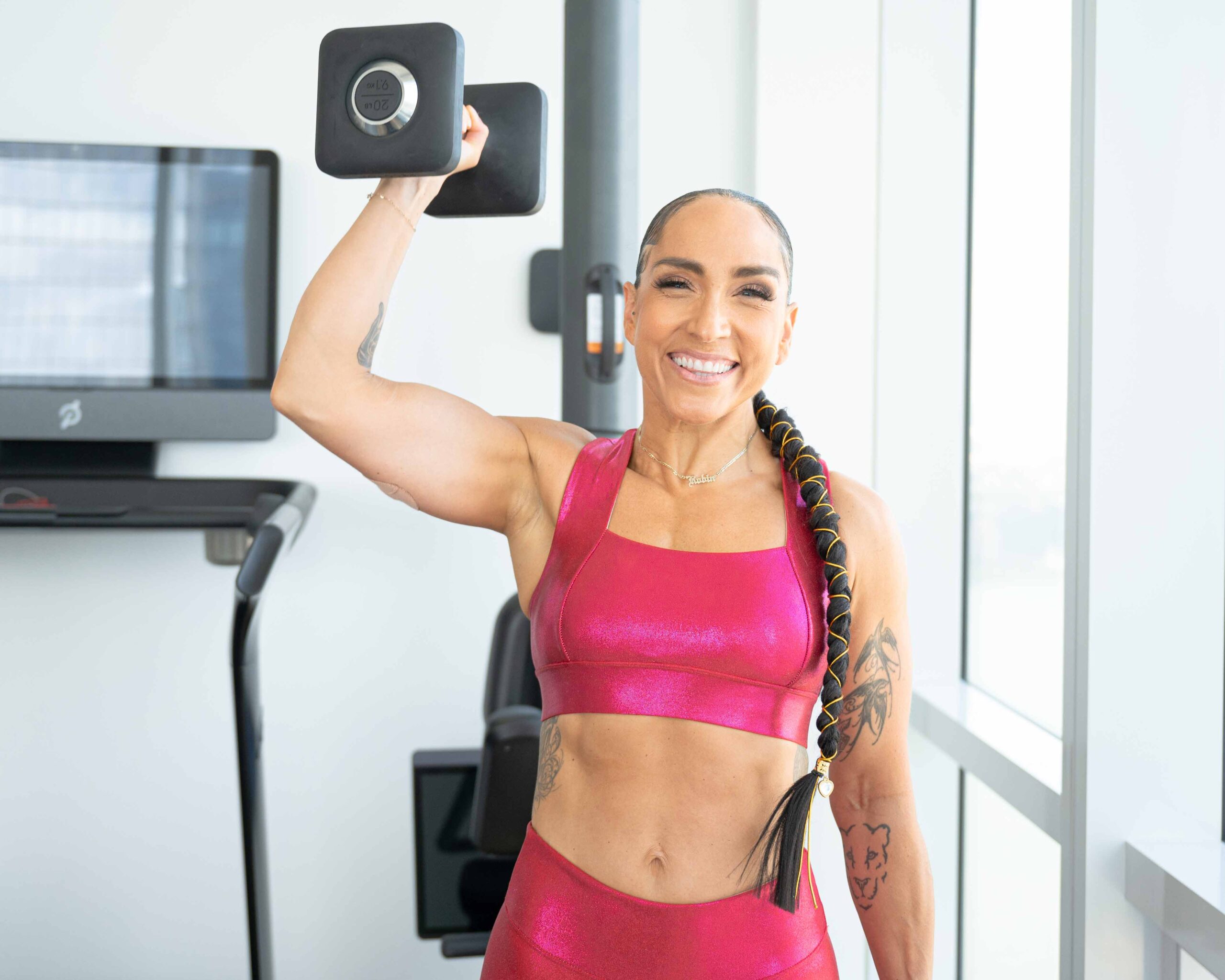 Head Peloton instructor Robin Arzón holds up a dumbbell in a gym and smiles, wearing a pink sports bra and leggings