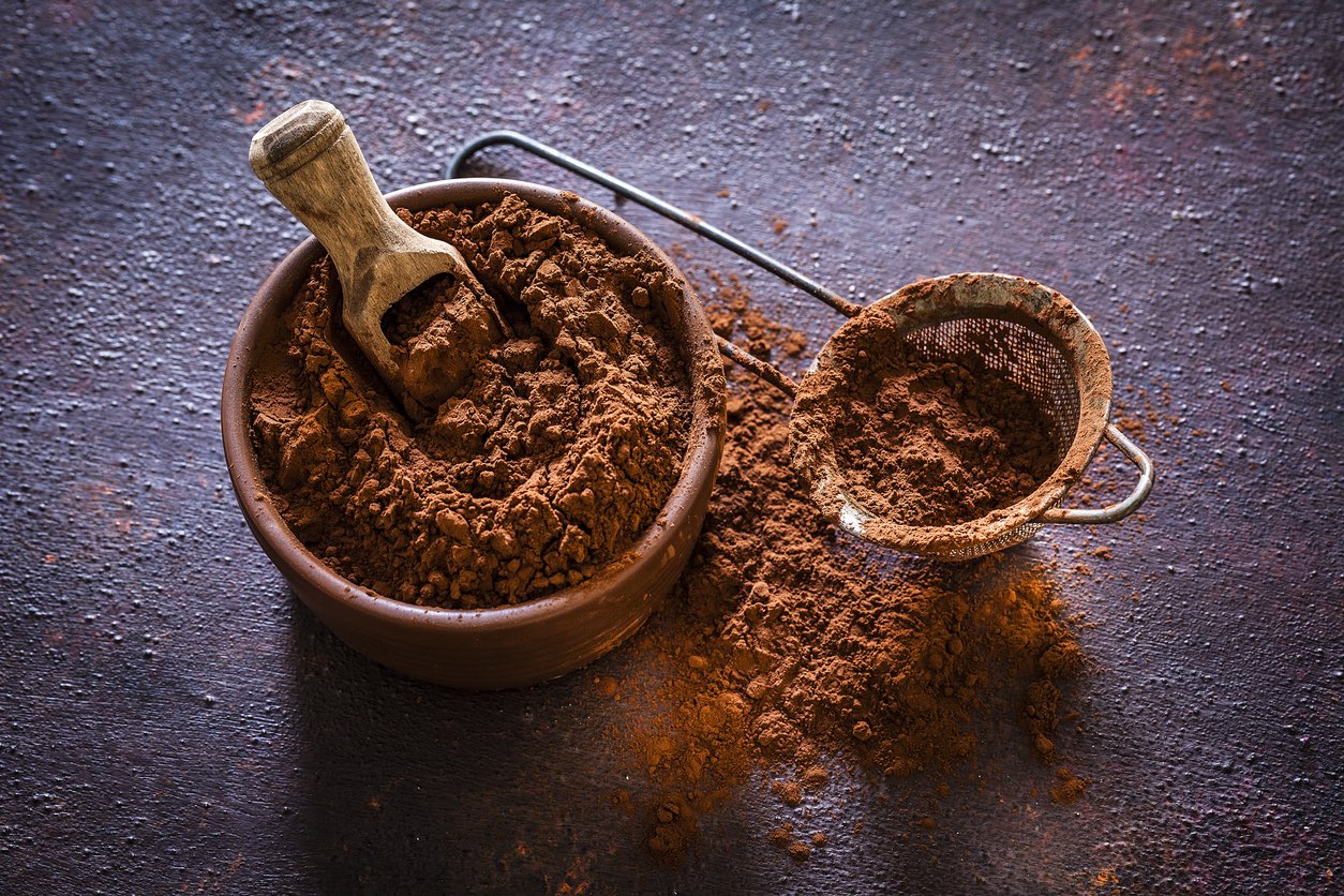 Brown bowl filled with cocoa powder shot on rustic table. Beside the bowl is a sieve also with cocoa powder.