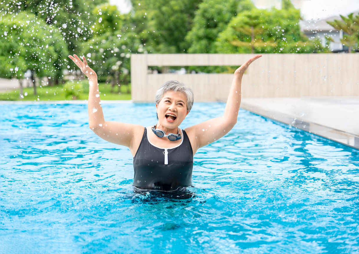 A senior woman stands in a pool outside looking joyous with her arms raised upward, wearing an athletic swimsuit and goggles around her neck
