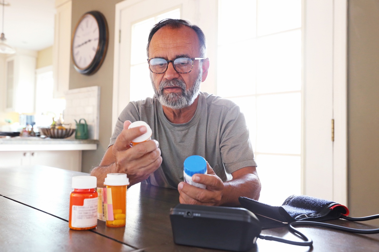 A senior citizen man sits at his dining room table sorting through bottles of prescription medications as sunlight filters in through the window behind him bathing the room with a soft glow of light. A blood pressure machine rests on the table in front of him.