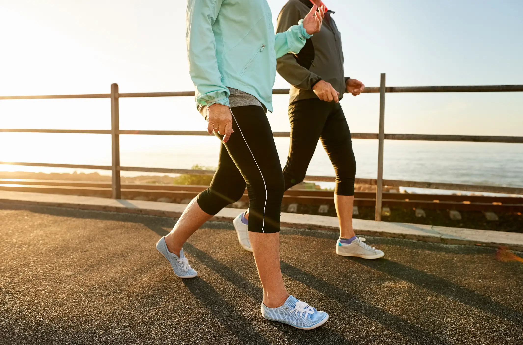 Two women shown from the shoulders down wearing windbreakers and leggings and sneakers, walking on a boardwalk