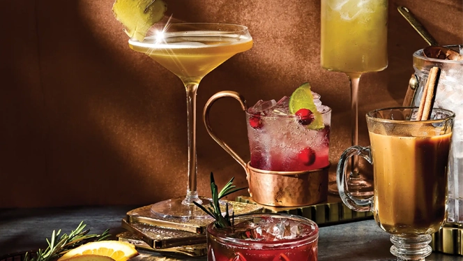 A styled holiday mocktail recipe spread featuring five drinks on a dark tabletop: a ginger-garnished coupe, a cranberry mule in a copper mug, a rosemary-cherry spritzer in a lowball glass, an herbal orange sparkler in a tall stemmed glass, and a warm spiced chai nog in a glass mug. Orange slices and rosemary sprigs sit in a small dish in the foreground, all set against a warm brown backdrop with dramatic light and shadow.