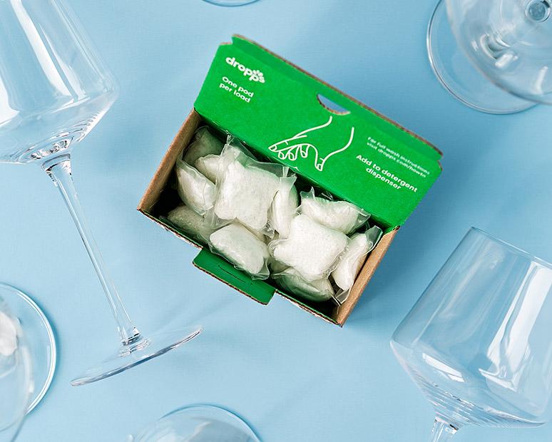DROPPS DISHWASHER PODS IN A BOX SURROUNDED BY WINE GLASSES AGAINST A BLUE BACKGROUND