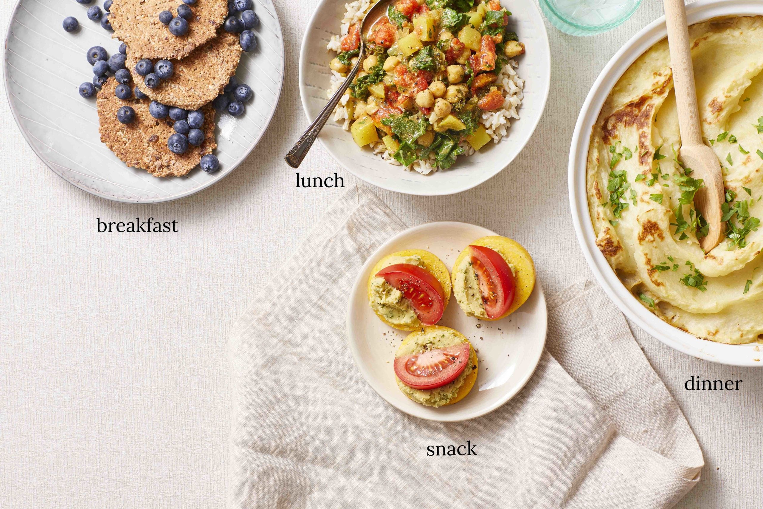 Day 6 of week vegan meal plan - white table with four dishes spread across it: Blueberry pancakes, chickpea casserole, a shepherd's pie, and polenta slices stacked with vegetables