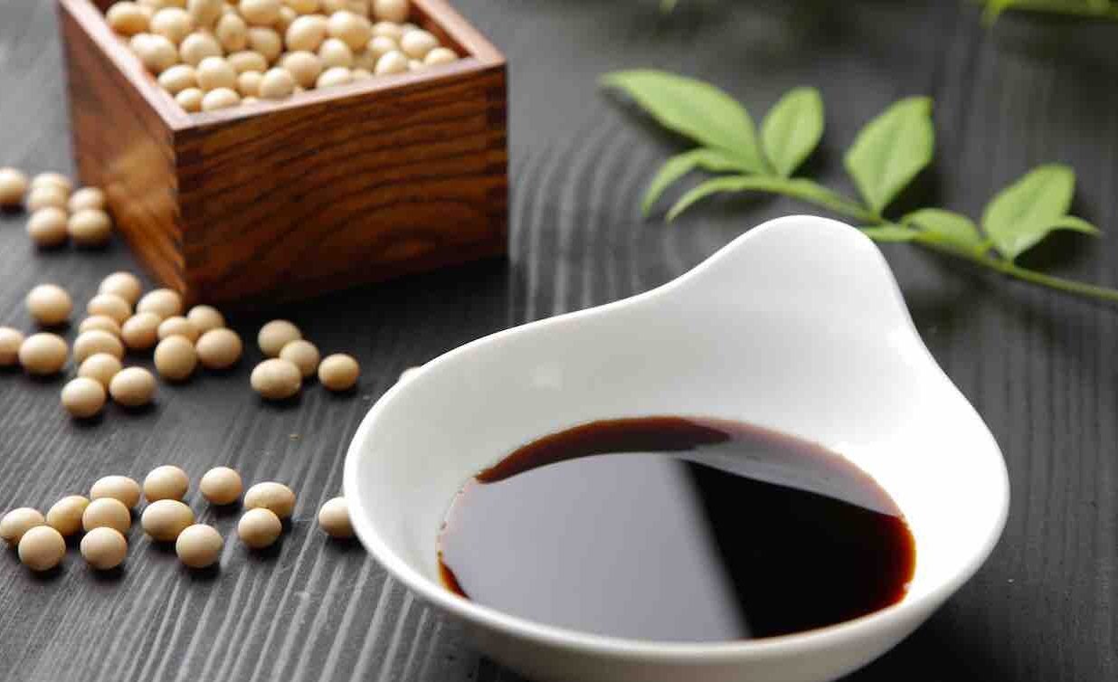 a small box of dried soy beans sits beside a small bowl of soy sauce, with some green leaves on the table behind it