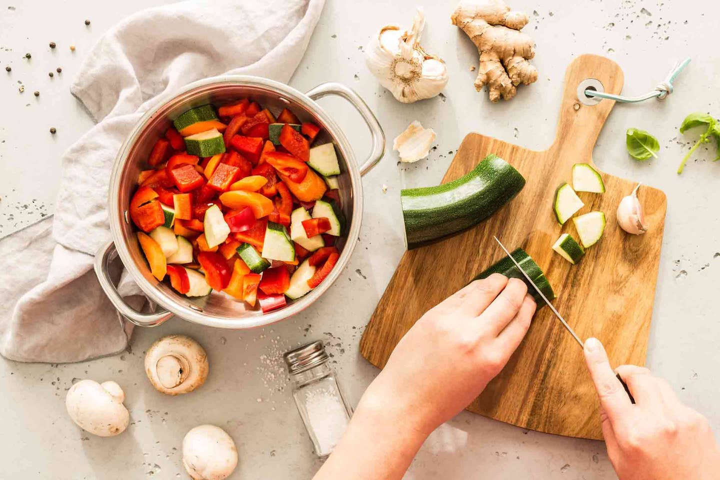 Woman prepping vegetables, slicing a zucchini on a cutting board, with a bowl of chopped bell peppers, tomatoes, zucchini to the side