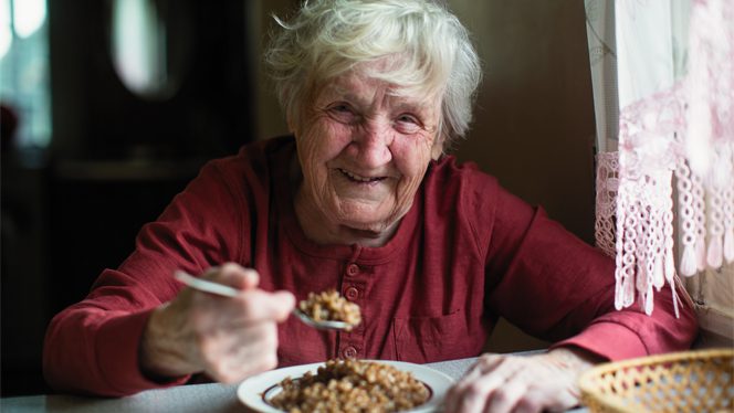 Old woman in red sweater eats a bowl of grains and smiles at the camera
