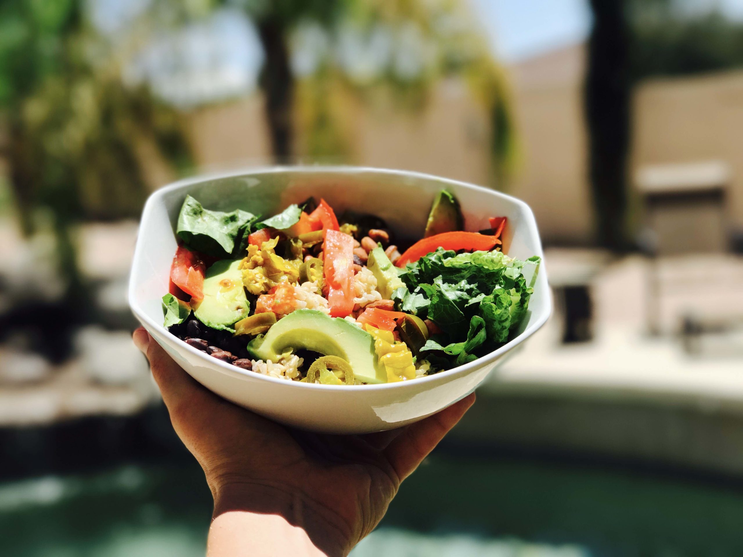 Vegan bodybuilder Robert Cheeke's hand holding up a plant-based burrito bowl, full of greens and grains, outside in the sunlight