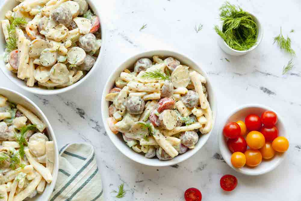 A healthy plant-based potato and artichoke salad shown in several white bowls, with a smaller bowl of tomatoes to the side