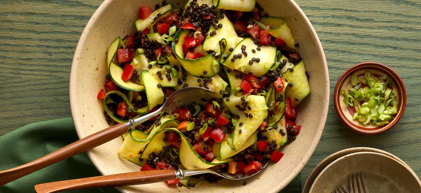 Ribboned zucchini salad with lentils and a creamy vegan dressing shown in a white ceramic bowl on a green wooden table