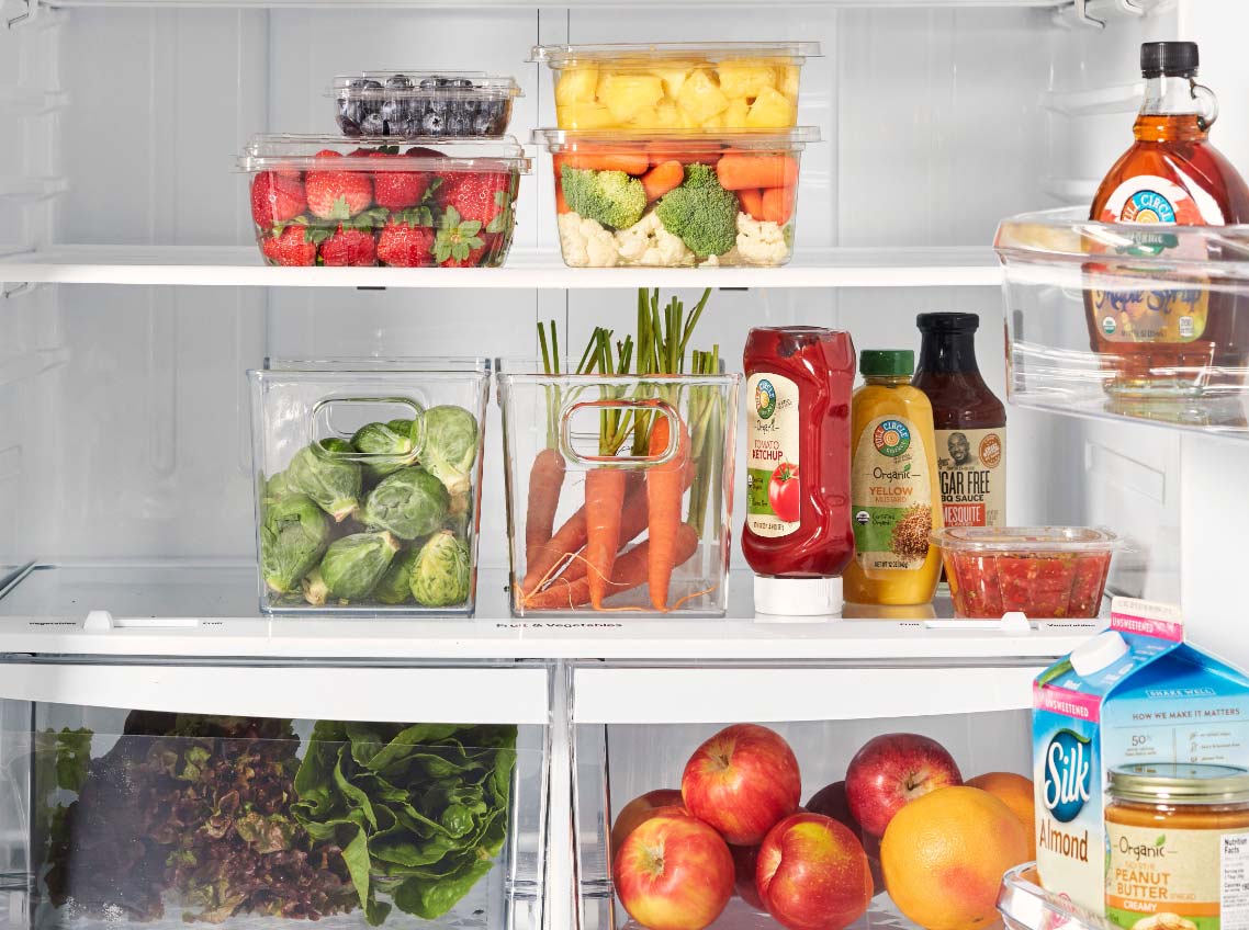 Healthy plant-based items in a refrigerator, including fresh fruit in drawers, pure maple syrup, and no-sugar-added ketchup