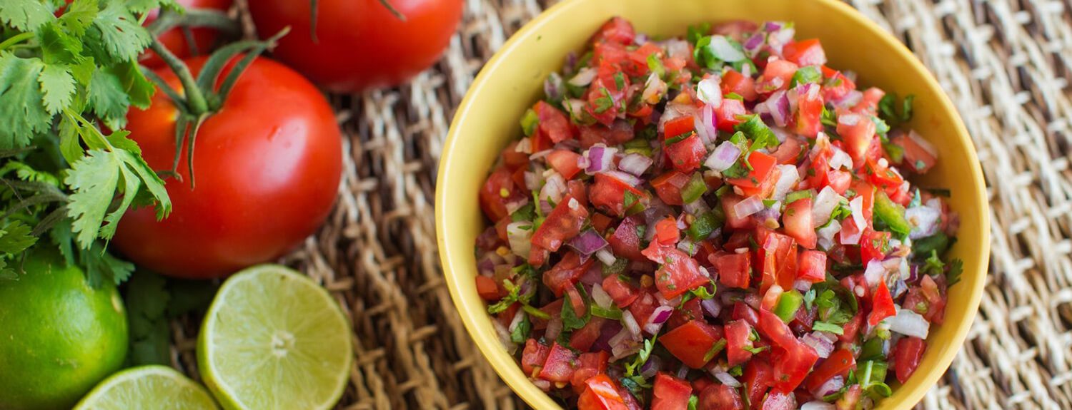 Fresh homemade tomato salsa in an yellow bowl beside fresh tomatoes, limes, and herbs