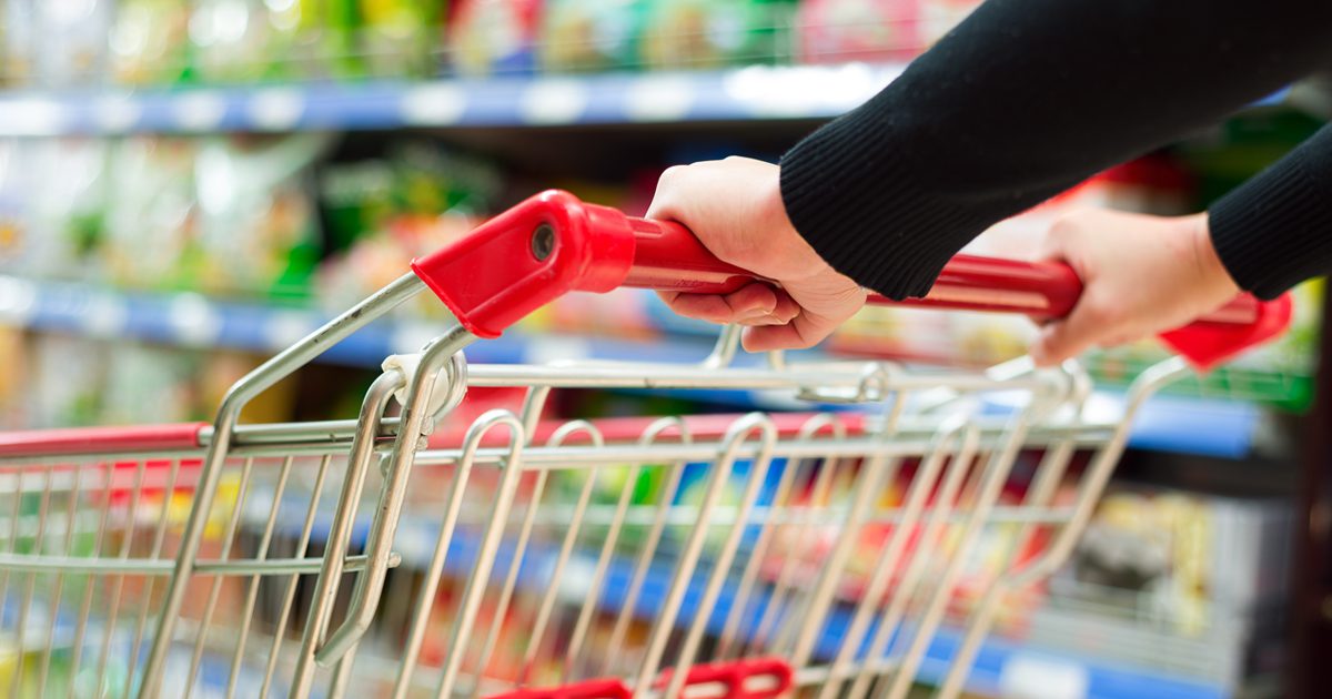 Hands on the red handle of a shopping cart, blurred grocery aisle in the background