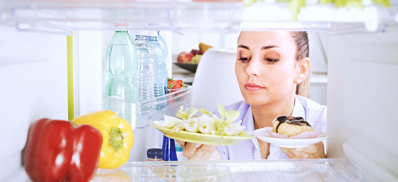 Photo taken from inside a refrigerator, with a woman looking in and examining a donut and vegetables