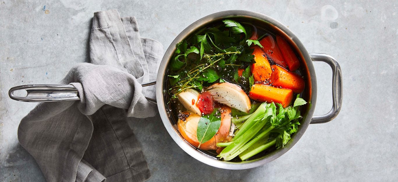 Vegetable Broth full of carrot chunks, celery, an onion wedge, and fresh herbs in a medium silver pot on a gray table with a gray cloth wrapped around the pot handle