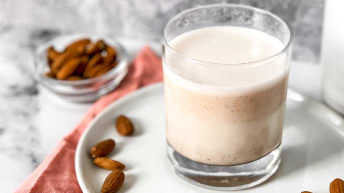 Almond milk in a clear glass on a white plate surrounded by whole almonds