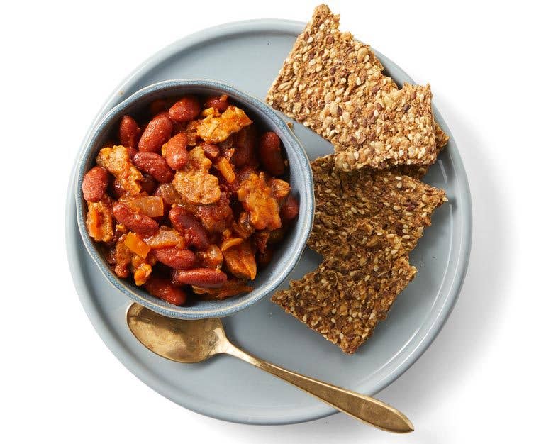 Breakfast beans in a small gray bowl on a plate with whole wheat crackers and a gold metal spoon