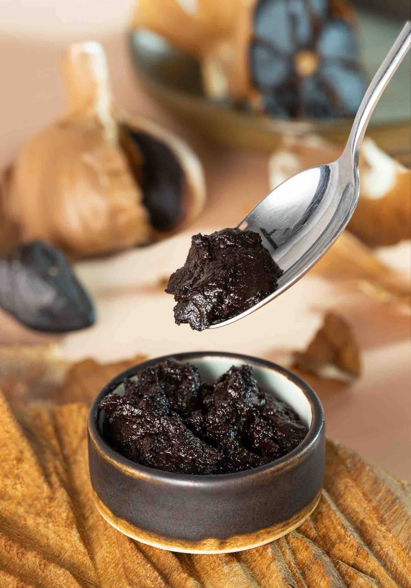 Spoon with black garlic paste over a small ramekin of black garlic paste, with a whole head of black garlic in the background