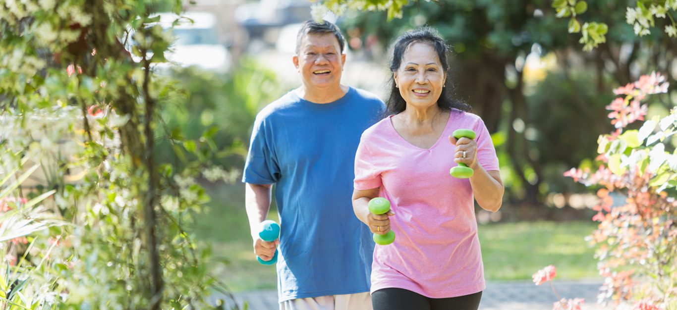 Senior Asian couple walk down a street holding hand weights and smiling