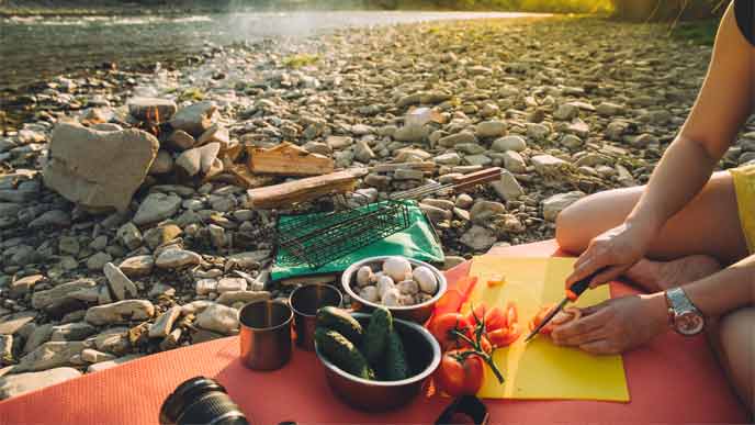 woman slicing veggies on a mat near a river with a campfire 