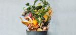 composting pail -- a metal bucket with fruit and vegetable scraps pouring artfully out, on a grayish white background